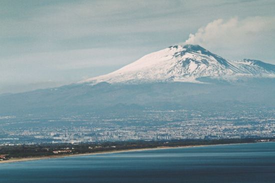 Schneebedeckter Ätna über der Küste bei Catania auf Sizilien mit ruhigem Meer im Vordergrund.