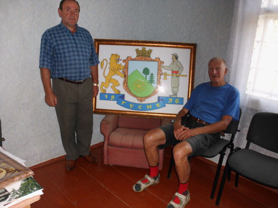 Im Gemeindebüro, einem einfachen Raum in einer Holzbaracke, erfahren wir mehr über das Schicksal dieser immerhin schon fast 450 Jahre bestehenden Siedlung. Man lebte von Landwirtschaft, Holzfällen und -bearbeitung. Bis heute hat sich daran nichts geändert. Vor dem Wappen von Huzne posiert der Bürgermeister. Der spitze grüne Hügel im Wappen symbolisiert den Berg Pikuj, den man von hier aus in 2 bis 3 Stunden besteigen kann.