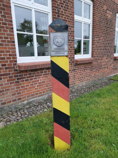 Die DDR-Grenzsäulen (auch DDR-Grenzpfähle genannt) waren Markierungen der Staatsgrenze der DDR und wurden erstmals im Herbst 1967 an der innerdeutschen Grenze aufgestellt.
Der Standort war anfangs etwa ein Meter hinter der Grenzlinie, später wurden sie etwa 5 bis 10 Meter auf DDR-Gebiet zurückversetzt. Grund waren Beschädigungen von westdeutscher Seite aus.
Die Grenzsäulen trugen unterhalb der Säulenspitze ein Schild mit dem Staatswappen der DDR und dem Staatsnamen. Anfangs handelte es sich noch um Plaketten aus Kunststoff, die mit einem mehrfarbigen Staatswappen bedruckt waren. Später wurden die Kunststoffplaketten gegen Reliefschilder aus Aluminiumguss ausgetauscht. Die Säulen waren rückseitig mit einer Nummernplakette versehen.
Die Säulen hatten einen Querschnitt von 22 cm im Quadrat und ragten je nach Standort 180 bis 210 cm aus dem Boden. Ihr Gewicht betrug etwa 400 kg. Aus der Spitze der Säule ragte ein 10 bis 15 cm langer Eisenstab. Dieser sollte verhindern, dass sich Vögel auf die Säule setzten und sie beschmutzten. Die Lackierung der Säulen verlief schräg von oben nach unten in den Farben schwarz - rot - gelbgold.
Insgesamt wurden 2735 Säulen von Pionierkräften der Grenztruppen aufgestellt. Die Säule mit der Nummer 1 stand am Priwall (Ostsee), Nr. 2735 im Dreiländereck BR Deutschland – DDR – Tschechoslowakei. Die Nummern waren in den Stabskarten der DDR-Grenztruppen aufgeführt.
(Quelle: Wikipedia)