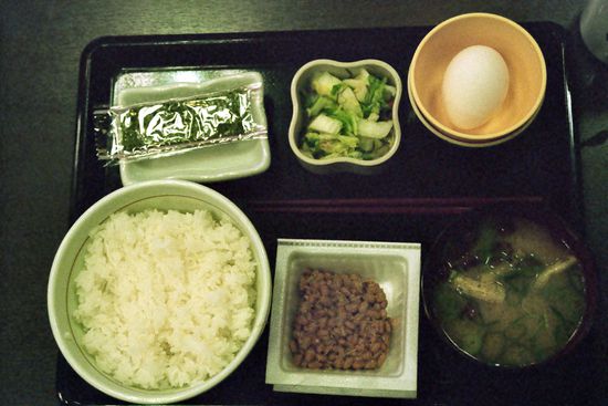 Frühstücksset (natto teishoku. Oben geröstete Algen, eingelegtes Gemüse, rohes Ei. Unten Klebreis ohne Salz, dermentierte Sojabohnen Natto, Miso-Suppe). Ei und Natto verquirlt über den Reis, dann mit den gerösteten Algen umwickelt, mit Stäbchen essen.