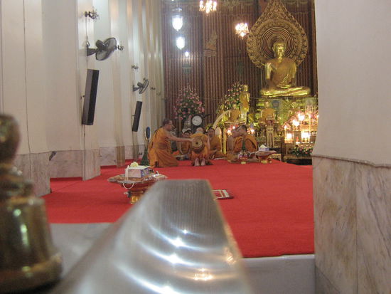 Der neue Mönch hat zum ersten Mal die Robe angelegt bekommen, der Kollege hilft ihm die Almosenschale richtig umzuschnallen.