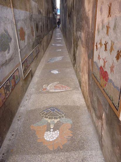 Die Schildkrötengasse, auf dem Boden sind Mosaike, an den Wänden verschiedene Arten von Schildkröten als Mosaik und ein Märchen über den Schutz von Meeresschildkröten in Mosaikform