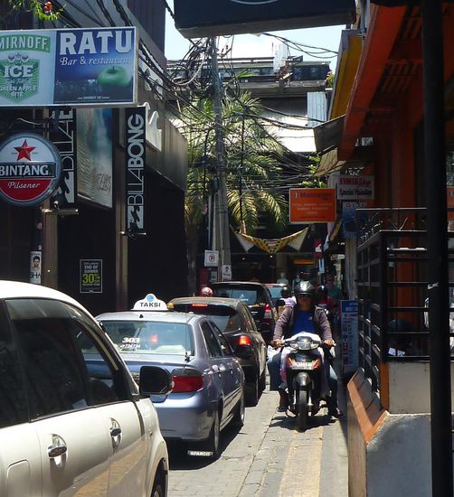 Typische Strasse in Kuta. Viele Autos, auf dem engen Einpersonengehweg kommen einem noch Mopeds entgegen,  wer lässt sich schon von Einbahnstrassen aufhalten! Allgemein wirksame Verkehrsregel : Es gibt keine und sollte es wirklich doch welche geben, werden sie höflich ignoriert ! Erstaunliches Fazit : Es funktioniert !!!