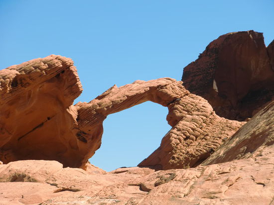 Valley of Fire - Der Name kommt bestimmt von den feuerroten Felsen!
Aber nach der Bekanntschaft mit dem Valley bin ich der Meinung, der Name kommt von der Gluthitze die dort herrschte!!!
Es wehte ein starker Wind! Gut für mich - so war die Hitze einigermaßenzu ertragen. Der Schweiß trocknete so schön im Wind!
Pech für Gregor - er haßt Wind und zieht schweißnasse T-Shirts auf der Haut vor!
Dieser schöne Steinbogen war kurz vor dem CP im Valley of Fire zu bewundern.
Der CP lag wunderschön zwischen den roten Felsen. Nachdem ich im Reiseführer gelesen habe daß diese Steine nachts die Wärme abstrahlen die sie tagsüber aufgeladen haben, war ich allerdings weniger begeistert von dem Platz.
Eintritt für den State Park 10$ pro Auto und für die Übernachtung auf dem CP hätten wir auch nur noch 10$ bezahlen müssen.
Wir übernachteten aber aus einem anderen Grund nicht hier: es war so heiß tagsüber, daß wir fast 4 L Wasser getrunken haben und nur noch etwas über 1 L Trinkwasser zur Verfügung hatten.
Also weiter - und einkaufen!
Wir hatten auf dem kurzen Weg von Las Vegas hierher keinen Supermarkt gefunden.
