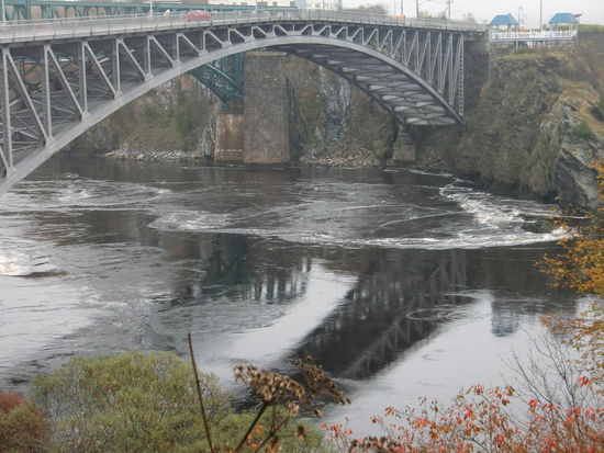 Die Reversing Falls am Morgen dem 18.10.: Da kommt die Flut von links von der Bay of Fundy und drückt das Wasser des Flusses zurück in sein Bett!
Wir haben direkt davor übernachtet! Hätte das Foto auch aus dem Auto heraus machen können!
Die Reversing Falls sind deshalb so einmalig da der Fluss eigentlich in Richtung Bay of Fundy fließt. Bei Ebbe fließt er auch in diese Richtung und bildet sogar einen kleinen Wasserfall dabei da über Felsen stürzt.
Kommt die Flut wird durch den gewaltigen Tidenhub der Bay of Fundy (bis 8,5 m) das Wasser des Flusses so stark nach hinten gedrückt daß er praktisch in die'  umgekehrte Richtung 'fließt'!
Bestimmt gibt es das nicht oft. Aber sooooo beeindruckend fand ich das jetzt auch nicht! Außerdem kann ich mir das gut vorstellen daß der Fluss wieder nach hinten in sein Bett gedrückt wird. Gregor fand das jedenfalls toll!