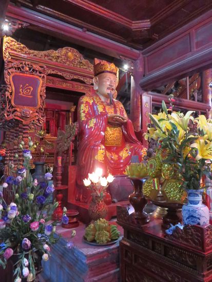 Auf einem Altar sitzt eine Statue von Konfuzius, der alte Gelehrte, rechts und links von ihm sitzen jeweils zwei Schüler, die ihm zuhören.