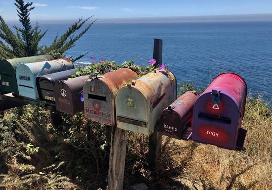 21 Briefkästen in einer Reihe. Der Postbote fährt per PKW vor - er hat sein Lenkrad rechts - und stopft aus dem Auto heraus die Post in die Kästen.