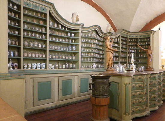 In den Mittelpunkt der Offizin gerückt zeigt sich der wichtigste Arbeitsplatz des Apothekers, der rezepturtisch, mit sechzig  sorgfältig beschrifteten Schubladen. Auf der eichenen Tischplatte wachen der antike Heilgott Aesculap und seineTochter Hygieia über die Arzneibereitung, in den Händen ihr typisches Attribut, den Stab mit der der Schlange haltend.