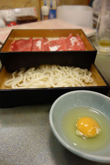 eine Platte mit Fleischscheiben, eine Schale mit dicken Odon-Nudeln und ein rohes Ei