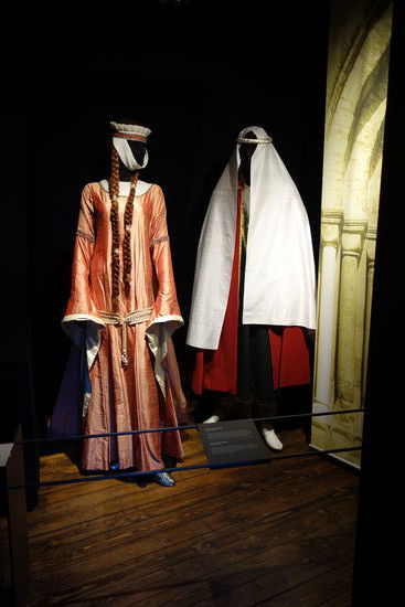 adelige Frauenkleidung - 12. Jh. 
seidenes Hemd, darüber ein Surcot mit Schleppe und Hängeärmeln aus antikfarbener Seide, an den Seitennähten geschnürt (eingenestelt) mit einem
aufwändig gearbeiteten Gürtel, dazu Schuhe aus Brokat, auf dem Kopf ein Schapel mit Gebende
adelige Männerkleidung - 12 Jh. 
langes Leinenhemd mit schmalen Ärmeln, darüber Suckenie (ärmelloser Überrock) mit echtem Pelzbesatz, halbkreisförmiger Mantel mit Tasselverschluss, Beinlinge mit Sohlen und auf dem Kopf einen gedrehten Kopfreifen