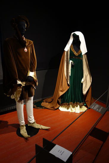 Kleidung einer deutschen Frau - um 1434
Cotte aus Seide, darüber ein Surkot mit Schleppe aus grünem Samt mit edelsteinbesetztem Samtgürtel und eine Houppelande mit Stehkragen perlenverziert aus reiner Wolle, auf dem Kopf eine Hörnerhaube mit Kruseler (Tuch mit gekräuselten Rand)
Männerkleidung -  15. Jh.
Tappert mit Zaddeln an Saum und Ärmeln, Ledergürtel, Beinlinge mit aufwendig gearbeiteten Schnabelschuhen, auf dem Kopf ein Barett mit Sendelbinde