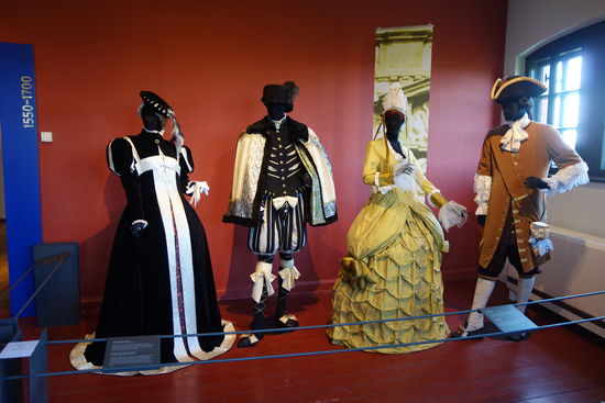 vlnr.
Kleidung einer Edelfrau - 16. Jh. (spanische Mode)
Reifrock mit Hüft- und Gesäßpolster, darüber ein Rock aus bestickter Seide mit Brokat und ein Umhang aus schwarzem Samt mit gepufften Ärmeln. Säume aus Brokat, mit Perlen bestickte Handschuhe, auf dem Kopf eine Calotte, darüber geschlitztes Barett aus schwarzem Samt mit Federn
Kleidung eines deutschen Höflings - 16. Jh. (spanische Mode)
Hemd aus Seidenleinen, darüber ein enges Wams mit gepufften Ärmeln und einer Pluderhose mit zu den Strumpfbändern passender großer Seidenschleife auf der Schamkapsel, darüber ein Umhang aus heller Wolle, gefüttert mit Brokat und mit schwarzem Pelz verbrämt, bestickte Strümpfe und schwarze Lederschuhe, Hut mit hohem steifen Kopf und geschwungenem Rand
Frauenkleidung - zweite Hälfte 17. Jh. (Barock)
Seidenunterhemd, sehr aufwendig gearbeitetes Mieder, Reifrock, darüber ein Kleid mit gepolsterten Hüften (Manteau) und überlangen Ärmeln, reich mit Perlen bestickt, auf dem Kopf eine Fontange mit weißer Spitze
Männerkleidung - um 1675 (Barock)
sehr aufwendig gearbeitetes Seidenhemd, lange Weste, geraffte Hose bis zum Knie, darüber ein Justaucorps (Jacke) aus mit Spitzenärmeln und einer Schärpe, Seidenstrümpfe, Schnallenschuhe, auf dem Kopf Dreispitz (Hut)