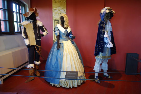 englischer Offizier - 17. Jh. (Frühbarock)
Seidenhemd mit Borte verziert, dazu ein Wams mit aufliegendem Spitzenkragen und hochgezogener Taille, halblange Hose, darüber ein Lederwams und Schwert mit Halterung, Stulpenstiefel, Stulpenstrümpfe, auf dem Kopf ein Kavaliershut mit Straußenfedern
Frauenkleidung - 17. Jh.
Reifrock mit Hüft- und Gesäßpolster, darüber ein Seidenkleid mit tiefem Ausschnitt und Umlegekragen, weiter, vorne offener Rock, Mieder mit Schneppentaille, auf dem Kopf ein edler Haarschmuck mit Perlen verziert
Männerkleidung aus der Zeit Ludwigs XIV.
sehr aufwendig gearbeitetes Seidenhemd mit Jabot, darüber ein boleroartiges kurzes Wams, sehr weite Pluderhose, darüber der Rock und ein reich bestickter Umhang aus dunkelblauem Samt, Seidenstrümpfe, Schuhe mit Ristlaschen, auf dem Kopf ein Hut mit Federn (Plumage)