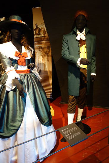 Kleidung einer Engländerin - um 1785
weiter Rock, darunter Reifrock mit Hüft- und Gesäßpolster, dazu eine Bluse mit herabhängenden Schleifen und einem sehr aufwendig gestickten Muff, Hut aus Seidentaft mit Federschmuck
Kleidung eines Engländers - um 1785
Hemd mit Ärmelvolants, dazu ein Gehrock mit hoch stehendem Kragen aus dunkelgrünem Tuch, eine Weste, und Hose mit Hosenlatz, Schuhe mit Absatz und Schnalle, Kopfschmuck war ein Samtband im langen Haar