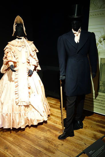 Damenmode um 1830 (Biedermeier)
zartfarbenes Kleid mit stark verbreiterten Schultern aus Moire Stoff, darunter ein steifer Unterrock, Mantille (Umhang), Sonnenschirm und Schute auf dem Kopf
Herrenmode um 1830 (Biedermeier)
Unterhemd, zartfarbenes Oberhemd aus Moire Stoff. Steghose mit roten Streifen, darüber ein taillierter Schoßrock, Lederschuhe, Stock und Zylinder