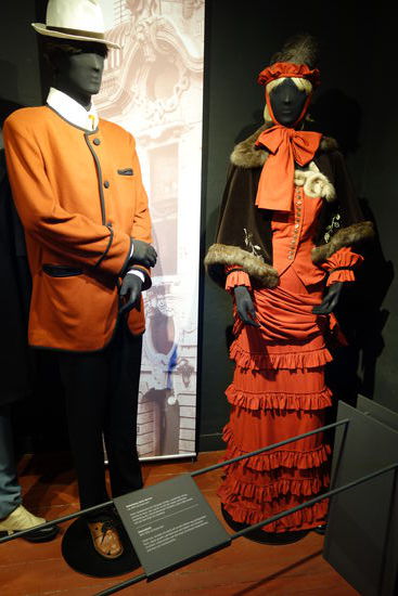 Bekleidung eines Herren - um 1870 (Gründerzeit)
helles Seidenhemd mit farbigem Schlips, zweiteiliger Anzug (schwarze Hose, rote Jacke), im Stil der neuen sachlichen Einstellung des industrialisierten Zeitalters, dazu Lederschuhe und auf dem Kopf ein Strohhut
Damenkleid - 1870-1890 (Gründerzeit)
Unterrock, darüber ein Kleid mit an den Hüften eng anliegendem Rock, üppig gerafft und überreich mit Falbeln und Schleifen versehen, Lederschuhe, auf dem Kopf ein kleines Federhütchen und dazu ein Sonnenschirm