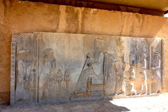 König Darius I. auf einem Thronrelief, die Figur links vom König hält eine Hand vor den Mund, eine Geste der Ehrerbietung