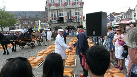 Der Käufer in weisser Kleidung und der Verkäufer beim feilschen um den Preis. Im Hintergrund das Rathaus von Gouda.