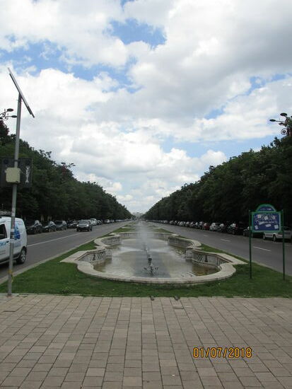 Fontänen in der Mitte der Straße. Fußgänger schlendern rechts und links durch eine parkartige Grünanlage