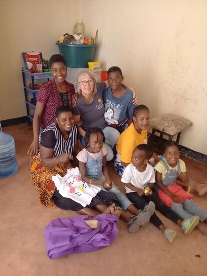 Familienfoto auf dem Boden. Im Hintergrund sieht man die Küchenutensilien. Im Nebenraum stehen zwei Betten für die ganze Familie. Es gibt noch einen Tisch und einen Fernseher. Viel mehr besitzen sie nicht.
