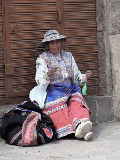 Diese Frau verdreht die gesponnene Wolle mit der Spindel, gemütlich vorm Haus sitzend.