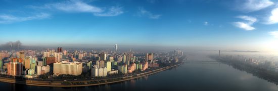 Panorama von Pjöngjang - von links nach rechts: Fabrikschlote, Koryo-Hotel (rotes Doppelgebäude), Ryugyŏng Hotel (große Pyramide), Juche-Turm (rechts vom Fluss)