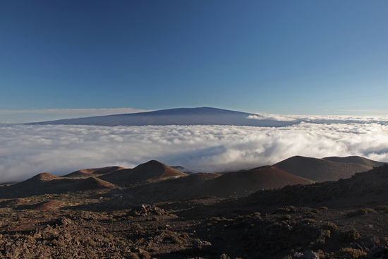Vom Abhang des Mauna Kea aus gesehen wirkt der Nachbarberg Mauna Loa wie ein riesiger Kuhfladen. Typisch für einen Schildvulkan. Mauna Loa ist einer der grössten und höchsten einzeln stehenden Berge des Sonnensystems, steigt er doch aus einer Meerestiefe von 5000m auf sagenhafte 4170m über dem Meer. Nicht nur, dass er damit über 9000m hoch ist, nein, er ist auch der an Volumen grösste Berg der Erde. Sein Rauminhalt beträgt 40000 Kubikkilometer. Eine abstrakte Zahl, die uns hier nicht weiterhilft. Anschaulicher ist folgendes Beispiel: könnte man den Mauna Loa aus dem Pazifik nehmen und nach Europa setzen, wäre er 9000m hoch und würde die gesamte Schweiz bedecken. Der Mauna Loa gehört mit dem Volcanoes National Park zum UNESCO Welterbe.
