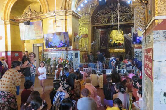 In der Mahamuni Pagode in Mandalay. Sie gehört neben der Shwedagon Pagode und dem Golden Rock zu den meist besuchten heiligen Stätten  in Myanmar.. Während die Männern im inneren Zirkel der Pagode sich der Buddha-Figur nähern und sie mit Goldplättchen versehen dürfen, bleibt den Frauen der äußere Raum zum Beten.  Viele Tonnen Gold in kleinen dünnen Goldplättchen verarbeitet wurden über Jahre auf die Figur geklebt, sodass die Statue bereits zur Unkenntlichkeit verändert wurde. 25 cm dick soll die Goldschicht aus 24 Karat Gold bereits sein. In eigens für die Goldplättchen eingerichteten Manufakturen werden die Goldblätter stundenlang in Handarbeit geklopft, damit der Spender sie dann käuflich erwerben und auf die Oberfläche des Buddhas anbringen kann. 
Unsere weiblichen Begleiterinnen scheinen sich mit der ungerechten Behandlung von Mann und Frau offenbar abgefunden zu haben. Auch wenn in der Verfassung die Gleichberechtigung verankert ist, so richtig durchgesetzt scheint sie sich noch nicht im täglichen Leben. Führungspositionen, egal ob in Staat oder Wirtschaft, werden lieber an Männer als an Frauen vergeben. Frauen hätten einfach noch zu viel tun mit der Familie, den Kindern, den Eltern, dem Haushalt usw. .