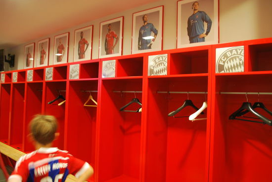 Etwa 10-jähriger FCB-Fan mit entsprechendem Trikot in der Kabine seiner Idole. Obwohl ich keine Hausfrau bin, fallen mir die Kleiderbügel auf: Da verdienen die Millionen, die Vereine küssen denen die Füße, sie spielen auf teuerstem Hybrid-Rasen, sie werden mitten in Berlin von Millionen Menschen gefeiert (teilweise) und von Helene Fischer geherzt, aber Kleiderbügel ??? Max. 2, davon mindestens einer aus Plastik, wenn nicht alle beide. Seeehr sympathisch !! Wie ich, schmeißen die Bayern-Spieler wohl auch alles auf den Boden.