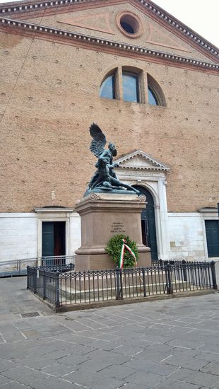Das Denkmal der Gefallenen vor der Kirche
Ein beflügelter Sieg, der einen Kriegsgefallenen stützt, ist ein Werk von Domenico Trentacoste. Die zwei bronzenen Statuen stehen auf einem Sockel aus rotem Granit, auf welchem man eine Gedenkschrift, über die Gefallenen des ersten Weltkrieges findet.