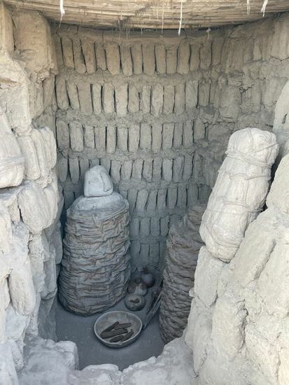 Ein Grab. Erkennbar die Totenmaske auf dem eingewickelten Leichnam und ein paar Essensbeilagen. Rechts scheint es sich um ein Baby zu handeln.