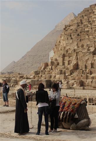 Ein Kameltreiber mit zwei Touristinnen und Kamel vor der Cheopspyramide; im Hintergrund die Pyramiden von Chephren und Mykerinos