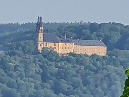 Nun also, es handelt sich um Kloster Banz, gar nicht mehr ominös und in der Längenausdehnung längs zum Maintal liegend. Vierzehnheiligen lag dagegen mit der schmalen Frontseite in Richtung Maintal.
Die auf 406 m Höhe liegende ehemalige Benediktinerabtei Kloster Banz nördlich von Bamberg und gegenüber der Basilika Vierzehnheiligen auf der westlichen Anhöhe des Maintals auf dem Banzberg gelegen, ist ein Gemeindeteil der Stadt 
Bad Staffellstein. Das Kloster ist im Besitz derf CSU-nahen Hanns-Seidel-Stiftung und dient als Tagungsstätte für rund 23.000 Gäste pro Jahr für rund 600 Veranstaltungen.
Um 1070 gegründet, musste das Kloster nach dem Dreißigjährigen Krieg neu gebaut werden und vertritt die Stilrichtung des Barock im Gegensatz zum Stil des Rokoko von Vierzehnheiligen.