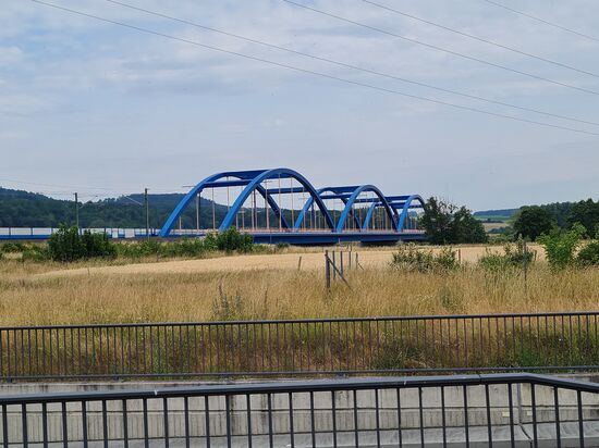 Zur Rechten meiner Fahrtrichtung überspannen drei auffällig in blau gehaltene Brückenbögen (Stabbogenbrücke) den Main bei km 416 (von der Mündung) in Nähe der Ortschaft Wiesen. Sie gehören zur Neubaustrecke Ebensfeld - Erfurt als Teil der Schnellfahrstrecke Nürnberg - Erfurt. und weiter nach Berlin.
Ja, es ist wieder soweit. Es gibt eine kleine Lektion vom Hobby-Eisenbahner:
Jeder Bogenteil hat 73 m Stützweite (insg. 219 m), ist 17,05 m hoch und wiegt 1000 Tonnen. 
Die Höchstgeschwindigkeit in  diesem Bereich beträgt 280 km/h.