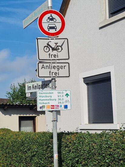 Kleiner Schilderwald in Bergrheinfeld:
(stinkende) Mopeds frei, gilt auch für normale Pedelecs, die bis 25 km/h 
vom E-Antrieb unterstützt werden.
Reine E-Bikes und Speed-Pedelecs, die jeweils 45 km/h erreichen können, sind von Haus aus auf die normale Straße verbannt, wobei auch Helmpflicht gilt.
Übliche Fahrradhelme reichen da nicht aus. Vom Gesetz aus wären sogar Motorradhelme nötig, aber die produzierende Industrie hat da einen gewissen Freiraum für besondere Ausführungen.
Für mich bis dato (beim Verfassen des Berichts Sept./Okt. '24) als altmodisch, konservativer Nicht-E-Radler bzw. Nur-Muskel-Kurbler sind diese Einschränkungen sowieso nicht relevant. Ich trage grundsätzlich schon seit vielen Jahren aus Überzeugung auch bei nur kurzen Radfahrten einen (nicht vorgeschriebenen) üblichen Fahrradhelm. Denn wenn die "Birne" Schaden nimmt, sind die Folgen oft drastisch schlimmer, als bei anderen Körperteilen. Da braucht auch niemand Hitzestau anführen, da die heutigen modernen Helme eine sehr gute Luftzirkulation ermöglichen.
Und als aufmerksamer Schilderleser, der auf zwei Radferntouren bereits in früheren Zeiten zweimal von Norddeutschland in die Heimat die Teilstrecke von Gemünden nach Würzburg am Main entlang zurückgelegt hat, ein drittes mal dieses Jahr 2024 in entgegengesetzter Richtung (siehe "Main-Radferntour 2024", zu einem späteren Zeitpunkt folgend), kann ich nur davon ausgehen, daß da die kürzeste Verbindung über die Prärie gemeint sein kann und nicht der Fahrweg über das von hier aus fast komplette Maindreieck entlang.
Egal, was soll's, weiter fahren ist angesagt...
.