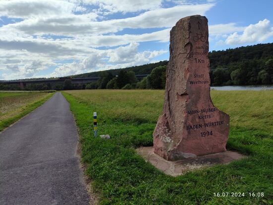 Noch etwas weiter entfernt rätselte ich an dem Felsmonument, ob es ein spezielles Denkmal sein könnte. Dabei handelte es sich um den mächtigen Grenzstein zum Wechsel des Bundeslandes Bayern, hier noch Main-Spessart-Kreis mit Kennzeichen MSP, nach Baden-Württemberg (Main-Tauber-Kreis TBB). Was auf dem Gestein nicht vermerkt ist: Der Mainradweg befindet sich ab hier anschließend im Regierungsbezirk Stuttgart. Im Hintergrund lichttechnisch etwas schlecht erkennbar die Mainbrücke der BAB 3 Nähe Wertheim aus Richtung Würzburg (links) nach Frankfurt/Main.