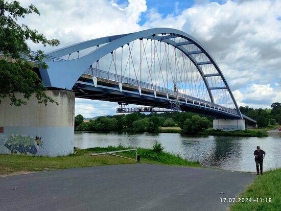 Die Roland-Schwing-Brücke von Niedernberg bei Großwallstadt (meine Seite) nach 
Sulzbach am Main. Insgesamt 318 m lang, hat der den Main überspannende Teil eine Stützweite von 150 m. Diese Stabbogenbrückenkonstruktion mit in Deutschland selten vorkommend gekreuzten Hängern vom Bogen zur Fahrbahn ist aber entlang  des Mains öfters anzutreffen, wie etwa von Grafenrheinfeld nach Bergrheinfeld (im Bericht der 2023er-Tour vorgestellt).
Die Ersatzübergangsstelle im Vordergrund für Amphibienfahrzeuge im Verteidigungsfall bzw. für Schwertransporte, die die Brücke nicht passieren können, wird auchv "Nato-Rampe" genannt und die Brücke auch NATO-Brücke.