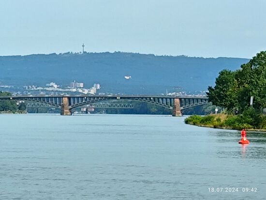Im Vordergrund die optisch ansprechende Theodor-Heuss-Brücke, der Konstruktion nach eine Stahlbogenbrücke mit Fachwerkbögen auf massiven Sandsteinpfeilern.
Sie wurde 1885 eröffnet und läßt auf ihr die B 40 auf einer Gesamtlänge von 475 m den Rhein mit dem frisch dazugewonnenen Wasser des Mains queren.
Im Hintergrund die Nordbrücke (nur für Bahnverkehr).