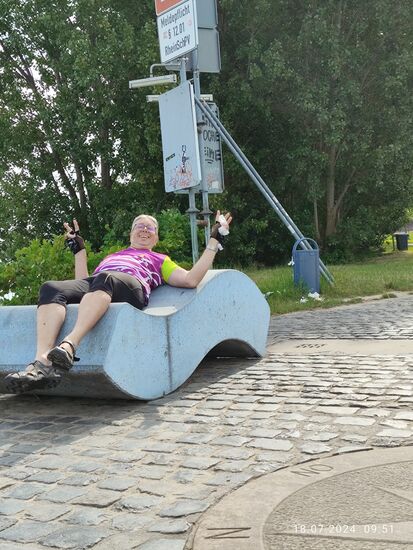 Die bläulichen Betonwellen bieten eine hervorragende Liegemöglichkeit. 
Hier noch ein Fernselfie mit dem selbstauslösenden Handy an einer Packtaschenfalte. Das V-Zeichen bitte ich wiederum nicht politisch zu deuten - es bedeutet für mich stets das gute Ende einer getätigten Aktion, wie z.B. an dieser Örtlichkeit das thematische Ende der Mainradtour der Jahre 2023 und 2024 !
