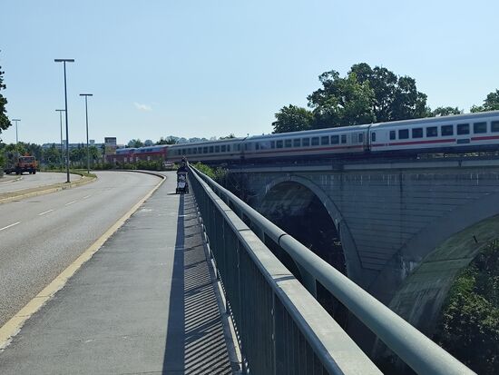 Wie bei meinen meisten Radreiseberichten so üblich, darf die Leserschaft wieder etwas Eisenbahnkunde "genießen" - hier in Form eines mittlerweile historischen Bildes. 
Ich konnte nämlich noch den IC 2084 "Nebelhorn" von Oberstdorf nach Hamburg-Altona auf der Illerbrücke ablichten, der in Augsburg Hbf nach Abkopplung der Diesellok-Doppeltraktion, hier links vorne, in den IC 2082 "Königssee" von Berchtesgaden nach Hamburg-Altona eingegliedert wurde und auch als IC 2082 "Königssee" weiterlief.
Leider wurden diese beiden Verbindungen zum Oktober 2025 eingestellt.
Zur Lok-Doppeltraktion noch folgender Hinweis:
Leider geht ja die Bildqualität beim Hochladen auf die Website etwas in die Knie, aber wenn man im rechten oberen Bildeck den schrägen, gegenläufigen Doppelpfeil anklickt, werden die Breitformatfotos vergrößert und wenn man weiß, wo man hinsehen muß, bemerkt man bei der Diesellok der Baureihe 218 parallel angeordnete Abgashutzen. Diese stehen hier für einen 12-Zylinder-Dieselmotor mit einem Hubraum von 9,56 Liter pro Zylinder und einer Motorleistung von 2800 PS.
Bei versetzt angeordneten Hutzen wäre es ein 16-Zyl.-Motor, aber mit weniger Hubraum und etwas geringerer Leistung. Bei der führenden Lok, auch Baureihe 218, ist die Hutzenanordnung leider durch die große Straßen-Hinweistafel auf die Gewerbebetriebe verdeckt.
Zum Fortfahren im Bericht übrigens das Kreuz im rechten, oberen Bildeck anklicken.