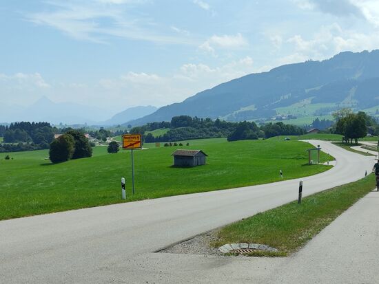 Am Ortsende von Oy (als nächste Ortschaft wird auf dem Ortsschild Nesselwang nach 3,5 km angekündigt) ergibt sich ein interessanter Fernblick:
In Bildmitte, etwa einen Finger breit vom linken Rand sticht trotz des Dunstes in der Ferne ein recht freistehender, pyramidenartiger Berggipfel hervor. Es ist der
2341 m ü. A. ("über Adria", wird nur in Österreich verwendet) hohe Thaneller, der in seiner direkten Umgebung der höchste Berg ist und einen freien Rundblick in alle Richtungen u.a. zum Zugspitzmassiv, die Ammergauer und Allgäuer Alpen ermöglicht. Links davon, aber bereits außerhalb des linken Bildrands, liegt mein Tagesziel Reutte in Tirol, das auf direktem Weg, immer schön entlang der Straße über Pfronten, Vils, Pinswang und Pflach überaus vorzeitig zu erreichen gewesen wäre. Aber warum schön und direkt entlang steuern, wenn bei diesem schönen Wetter ein schöner Umweg (der Umweg ist das Ziel !) eingeplant war.
Für mich war also angesagt, hinter dem linken Fuße des dunklen Höhenzugs in Bildmitte nach rechts abzudrehen und das Tannheimer Tal anzusteuern.