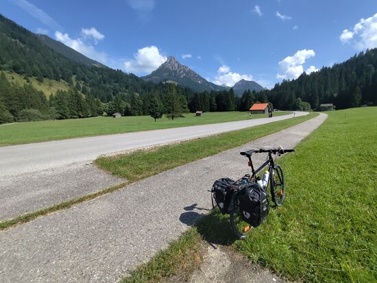 Ein erstes gutes Stück Strecke kann man zum größten Teil auf einem gut ausgebauten, teils welligen Kiesweg auf der gegenüberliegenden Seite der Steinacher Achen abgekoppelt vom Straßenverkehr bewältigen, dann beginnt noch weit vor der Landesgrenze von Bayern nach Tirol ein sehr gut ausgebauter 
Radler-Highway, gelegentlich muß man beim Überfahren von Weiderosten etwas Vorsicht walten lassen. 
Nicht nur an diesem Tag, auch anhand vieler anderer Erfahrungsberichte über lange Zeit sollte noch angeführt werden, daß der Radweg aufgrund der recht niedrigen Verkehrsdichte und des praktisch nicht vorhandenen Schwerverkehrs eigentlich überflüssig ist. Trotzdem ist dessen Vorhandensein natürlich sehr lobenswert, um jeglichen direkten Kontakt zwischen Auto-Mafia und Radler-Camorra zu vermeiden (beide haben ja so ihre Negativerscheinungen im rollenden Miteinander ...)
Bei diesem Zwischenhalt war ich mit dem Kalibrieren des Smartphone-Kompasses beschäftigt, wobei man das Handy einige Zeit lang in großen Achter-Schleifen vor dem Körper bewegt. Da kommt doch ein frecher Motorradbiker, der auch eine kleine Pause einlegte, auf mich zu und fragte mich, ob ich denn Schmetterlinge mit dem Handy fangen würde.
So ein Scherzbold ... 
Aber während der kurzen Pause haben wir uns bestens unterhalten.