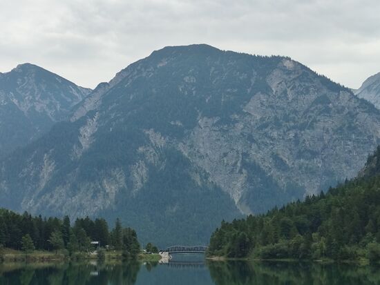 Hier ein Zoombild vom Beginn des (Kleinen) Plansees, der die Engstelle zum Beginn des (eigentllichen) Plansees und die schon angesprochene Seespitzbrücke zeigt.
Überquert man sie am Straßenparkplatz von links nach rechts, kann man hinter dem Waldriegel teils direkt etwas oberhalb vom Ufer an einem gut befahrbaren Trail entlang eines ca. 300 m langen Verbindungskanals zum Heiterwanger See gelangen, der bald hinter dem rechten Bildrand erscheint. 
Er hat eine Fläche von 1,37 km², eine Länge von 2,2 km, ist 820 m breit, hat ein Volumen von 55,3 Mio. m³ und eine max. Tiefe von 60 m. 
Eine der höchstgelegenen kommerziellen Schifffahrtslinien Österreichs bietet von Ende Mai bis in den Herbst hinein Rundfahrten auf dem Plansee und Heiterwanger See an, allerdings war zumindest an diesem Augusttag keine Flottenbewegung wahrnehmbar.