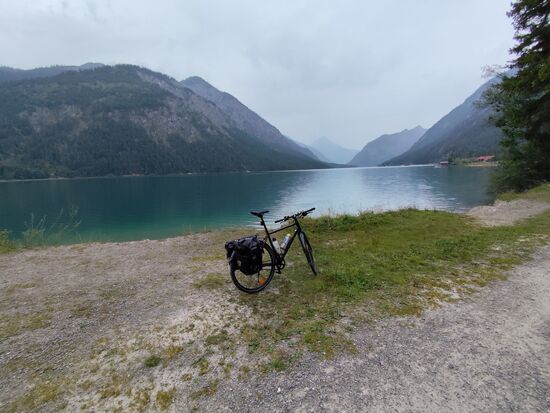 Etwas Entspannung ist also angesagt und bei eingeklickter Schuhsohle war die ca. hälftige Ablösung ja am Pedal fixiert. Im hinteren Bildbereich führt dann die Planseestraße in nördliche Richtung zum Bergrücken ganz im Hintergrund. Dieser erst noch tiroler und später bayrische Teil wird dann als Ammergebirge mit dem Ammerwald im Herzen dieses Gebirges (das größte Naturschutzgebiet Bayerns) bezeichnet.