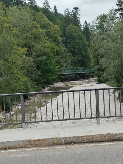 In Flußrichtung sieht man die alte Stahlfachwerkbrücke aus dem Jahre 1894, die ebenfalls vom bzw. zum Schloß Linderhof führt. Sie wurde nach zwei von einem Hochwasser zerstörten Holzbrücken aus den 1870er Jahren durch eine solidere Eisenkonstruktion ersetzt.
