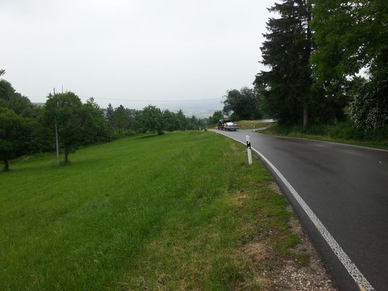 Auf der "Paßhöhe" des steilen Anstiegs (in direkter Nähe zum Wasserberg (751 m)): Blick zurück in den Westen. Bedingt durch das trübe Regenwetter sind die Niederungen leider nur schemenhaft als etwas grauerer Bereich oberhalb der nach unten endenden Straße erkennbar.