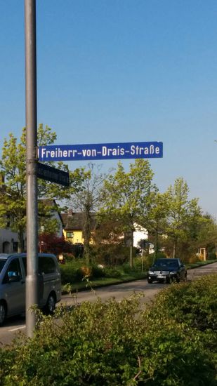 An diesem Straßenabzweig in Edingen-Neckarhausen, das durch den Verlauf des Neckars zum nördlich gelegenen Ladenburg abgegrenzt wird, darf eine spezielle Huldigung natürlich nicht ausbleiben:
Karl Freiherr von Drais, ein Universal-Erfinder, hat mit seiner wichtigsten Erfindung, der zweirädrigen Laufmaschine (Ur-Fahrrad) am 12. Juni 1817, also vor knapp 199 Jahren, seine erste Ausfahrt über 14 km getätigt - ohne Packtasche...
