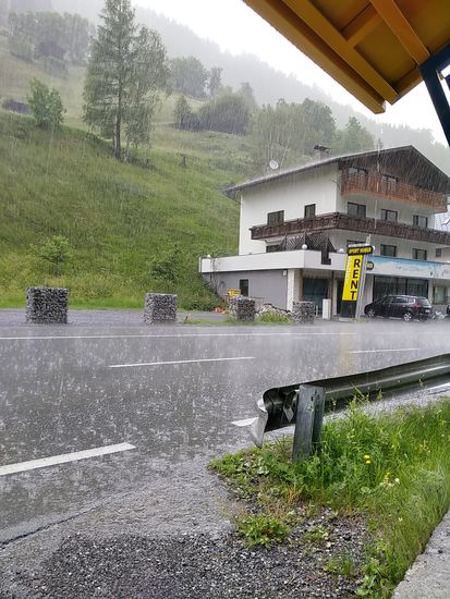 Es muß kurz vor der Ortschaft Kappl gewesen sein, als die Luftfeuchtigkeit drastisch überhand nahm. Vielleicht 100 m vorher zog ich bei einsetzendem Regen meine Schutzkleidung über, traute der Lage jedoch irgendwie instinktiv nicht so recht. Nur kurze Zeit in einem Bushäuschen untergeschlüpft (rechts oben ist ein Teil vom Dach erkennbar), setzte sintflutartiger Regen ein. Da wäre auch ein Weiterfahren mit Regenkleidung nicht nur unangenehm, sondern auch aufgrund eines fehlenden Radwegs und der schlechten Sicht für Autofahrer nicht gerade ungefährlich gewesen. Trotzdem fuhren andere Radler in meine Richtung weiter, ohne sich unterzustellen. Dabei war der Sturzregen nach max. 10 min in einen abklingenden Regen übergegangen.
Nur zu gut, daß die Höhepunkte des Tages, die auch das Kernthema der gesamten geplanten Radferntour waren, noch bei Top-Wetter ihre überaus bewegende Wirkung auf mich entfalten konnten !
DANKE, WETTERGOTT !!!