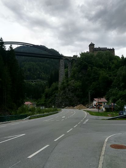 Es ist zwischenzeitlich 17:17 Uhr, die Straße schon seit einigen km abgetrocknet, auch die Regenkleidung, sodass ich diese hier, in einer Bushaltebucht pausierend, wieder in die Gepäcktasche verstauen konnte.
Linkerhand der Straße fließt die das Paznauntal entwässernde Trisanna, die bei Galtür durch den Zusammenfluss von Vermuntbach und dem Jamtalbach ihren Ursprung hat.
Gleich hinter dem Kraftwerk Wiesberg, dem Gebäude in Bildmitte links, vereinigt sich von links der 42 km lange Fluß Rosanna, der nördlich von Wirl bei Galtür seinen Ursprung hat und über St. Anton am Arlberg und über das Stanzertal hierher gelangt, mit der etwa 31 km langen Trisanna zur Sanna, die Ihrerseits nach nur ca. 7 km in Landeck in den Inn mündet.
An Bauwerken fällt an dieser Stelle die optisch imposante Trisannabrücke mit 230 m Länge und einer Höhe von 87 m auf, über die der Eisenbahnverkehr der vielbefahrenen Arlbergstrecke zwischen Landeck und Bludenz, wo ich gestern ins Montafon gewechselt bin, stattfindet. 
Auf diesem Bild geht's auf der Brücke rechts in Richtung Landeck an Schloss Wiesberg vorbei, einer Burg aus etwa dem 13. Jahrhundert, die sich heute in Privatbesitz befindet. Auf optisch halber Höhe der Trisannabrücke sieht man im Hintergrund ein Stück der Arlberg-Bundesstraße, die Arlberg-Schnellstraße verläuft noch weiter nördlich im weiteren Bereich hinter dem Bergrücken.
Grundsätzlich ist zu sagen, daß die Arlberg-Strecke mit Straße und Bahn die Hauptverkehrsader zwischen Tirol und Vorarlberg darstellt und die von mir beradelte Silvrettastraße den touristischen Ableger.