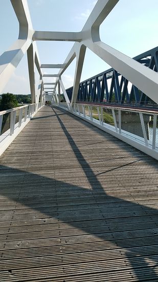 Die direkt daneben liegende, in hellgrau gehaltene Fußgängerbrücke oder auch Donausteg Deggendorf (einen offiziellen Namen gibt es nicht) wurde in 2014 eröffnet und wurde anstelle der in 2010 abgetragenen alten Eisenbahnbrücke errichtet. Mit 456 m ist sie 10 m kürzer als die stromaufwärts direkt nebenan liegende Neue Eisenbahnbrücke, die im Bild oben beschrieben ist und zählt zu den längsten Geh- und Radwegbrücken in ganz Europa.